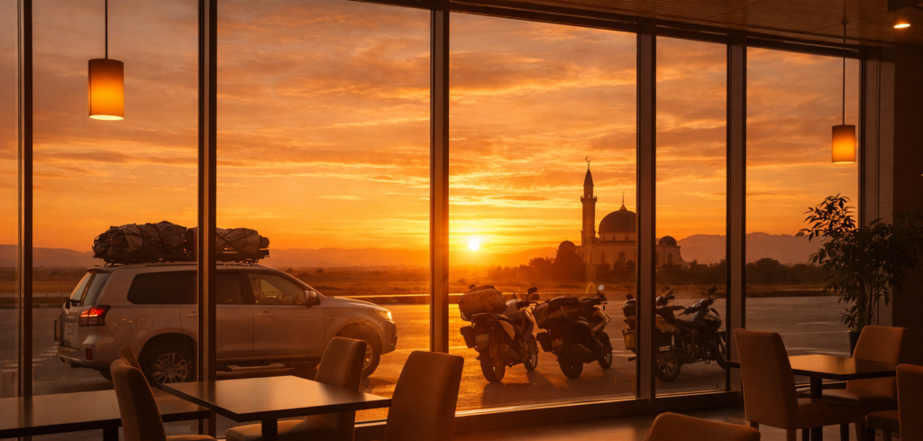 Interior rest area saat golden hour di bulan ramadhan dengan cahaya senja masuk dari jendela besar, terlihat mobil dan motor mudik terparkir serta siluet masjid di kejauhan.