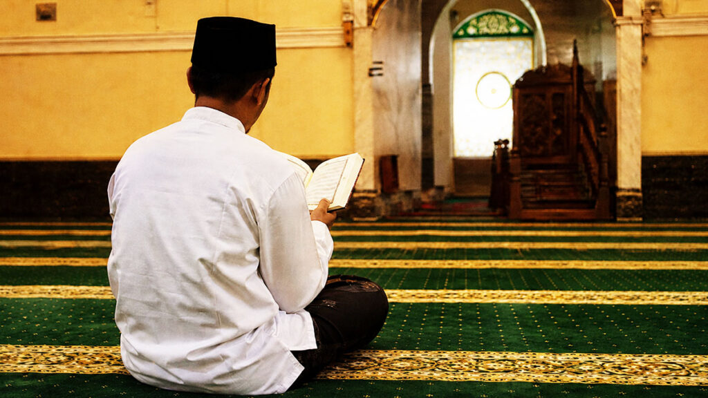 Seorang pria memakai baju koko putih dan peci hitam membaca Al-Qur'an di dalam masjid pada 10 hari pertama Dzulhijjah