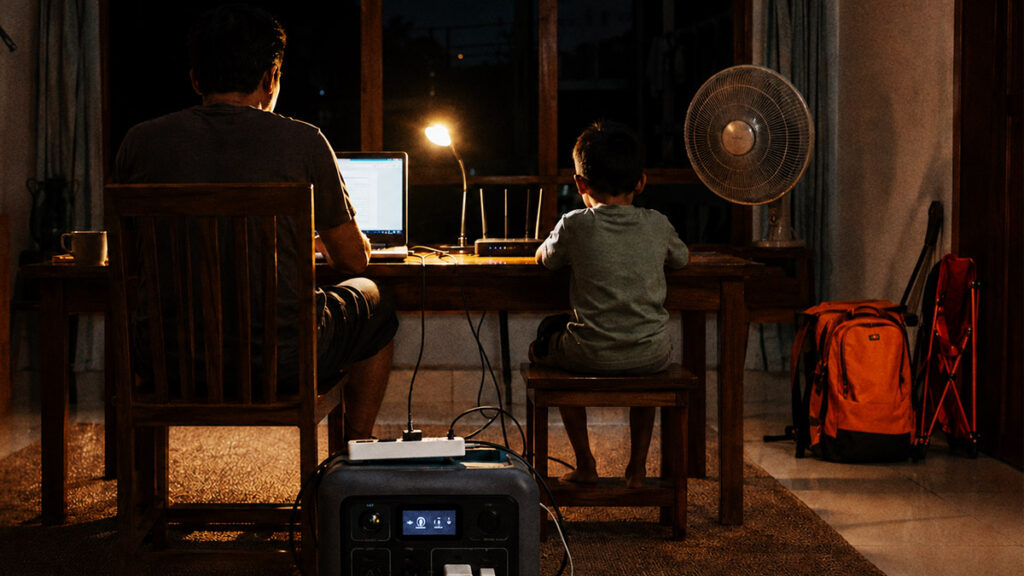 Ayah dan anak menggunakan power station untuk menyalakan laptop, WiFi, lampu, dan kipas saat listrik padam.
