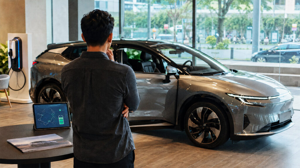 Calon pembeli melihat mobil listrik di showroom sambil mempertimbangkan klaim range dan standar jarak tempuh EV.