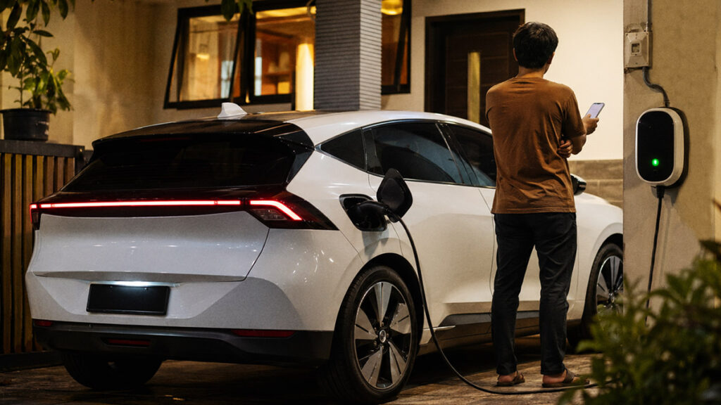 Mobil listrik sedang dicas di rumah pada malam hari dengan pemilik melihat ponsel di dekat meteran listrik